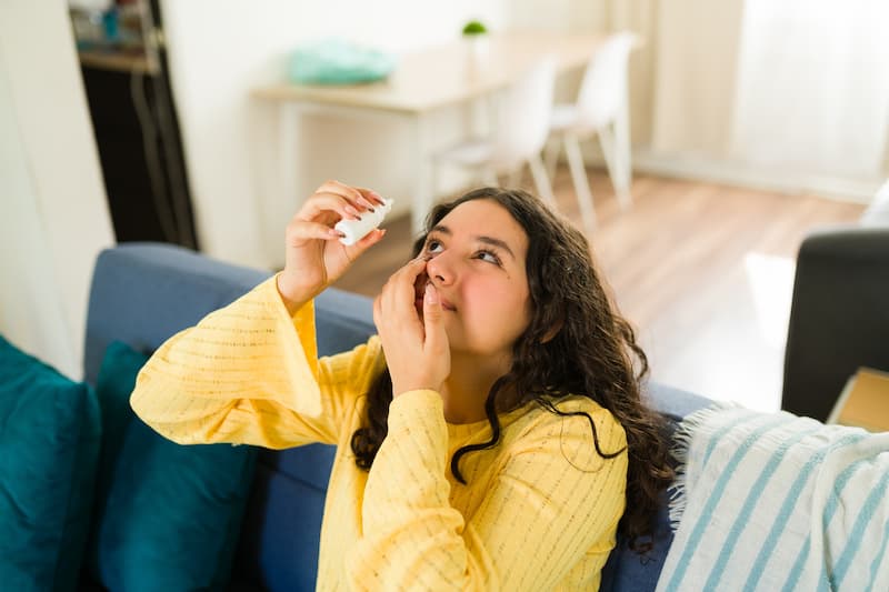 Teenager applying eye drops while sitting on a comfortable sofa